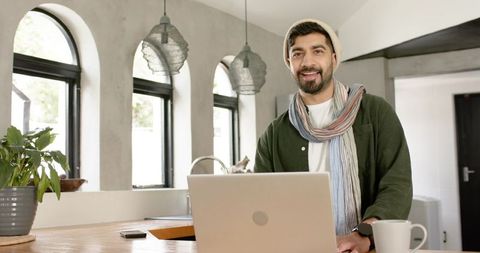 Indian man working remotely in kitchen with laptop, beanie and scarf, smiling