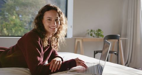 Smiling Woman Relaxing with Laptop on Cozy Bed Near Sunlit Window