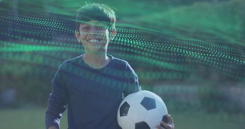 Smiling Indian boy holding soccer ball on park field with green digital wave overlay
