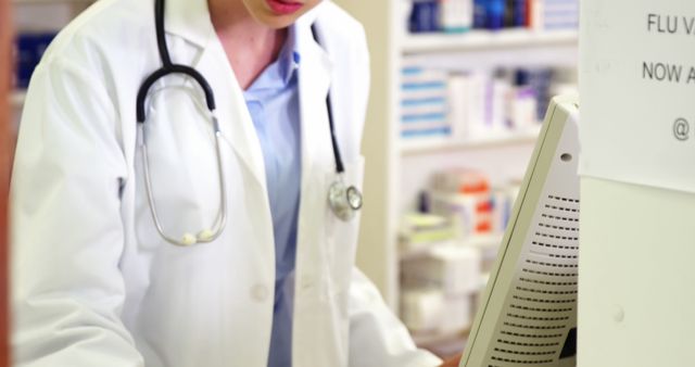 Healthcare professional in a white coat using computer in pharmacy or clinic. Person wearing stethoscope, indicating medical profession. Ideal for illustrating healthcare technology, pharmacy management, or medical professionals at work. Useful for websites, brochures, and articles focusing on medical services, pharmacy practice, or healthcare systems.