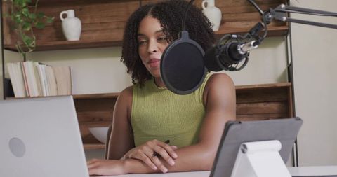 African american woman recording podcast at home office with laptop, tablet and condenser mic