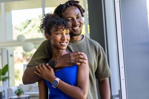 African american couple embracing by glass door wearing smartwatches in sunlit modern home
