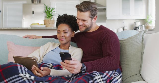 Image of happy diverse couple embracing and using electronic devices on sofa. Love, relationship and spending quality time together at home.