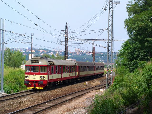 A red and white passenger train is traveling on curved railway tracks through a green and hilly countryside. Surrounding vegetation is lush, and electric poles and lines run alongside the tracks. The image conveys themes of travel, transportation, and rural journeys. It is ideal for use in articles, advertisements, or promotional material related to travel, train journeys, or nature exploration.