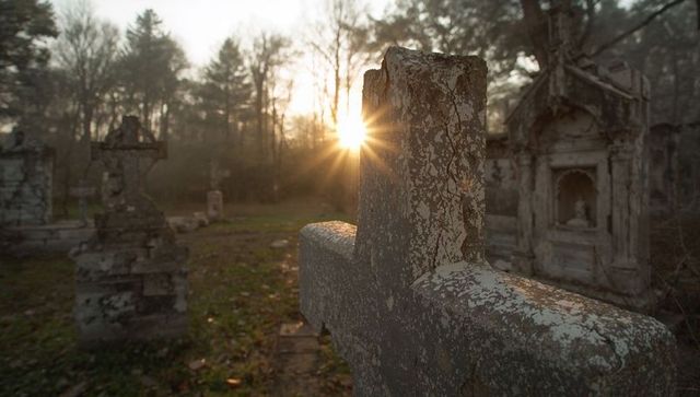 Lichen-covered stone cross at sunrise in woodland cemetery with sunburst