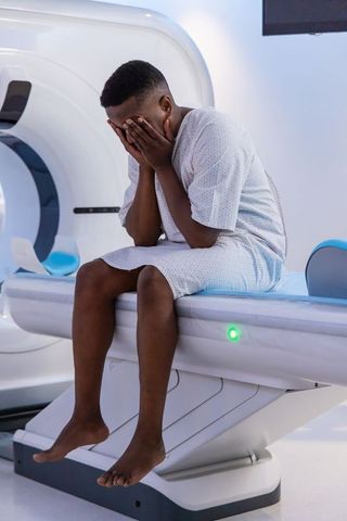 Male patient sitting on ct scanner table covering face wearing hospital gown