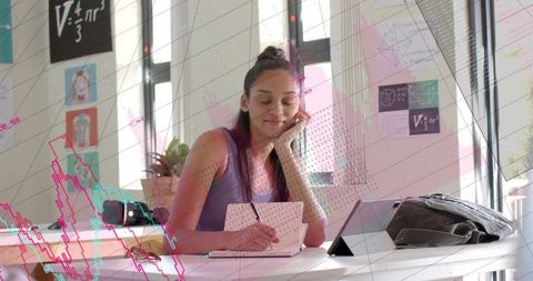 Focused student writing journal entries at sunlit desk with tablet and backpack
