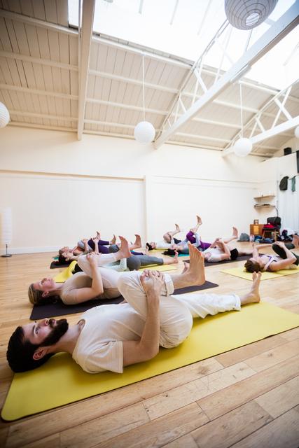 Group of people lying on yoga mats in a bright, spacious fitness studio, performing stretching exercises. Ideal for promoting fitness classes, wellness programs, and healthy lifestyle campaigns. Useful for illustrating teamwork, physical activity, and relaxation in a gym or wellness center.