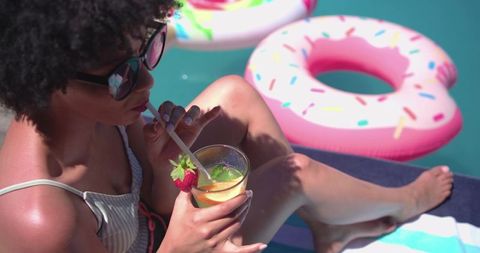 African american woman sipping cocktail poolside, lounging on towel with pink donut float