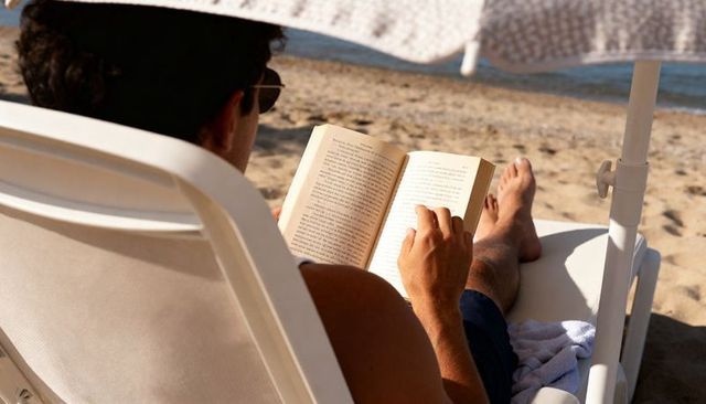Man relaxing on beach chair reading paperback under umbrella at sunny seaside
