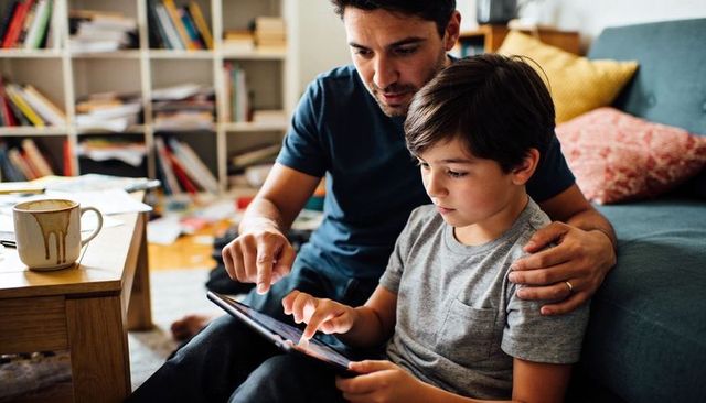 Father guiding son on tablet on living room floor, parent-child digital learning