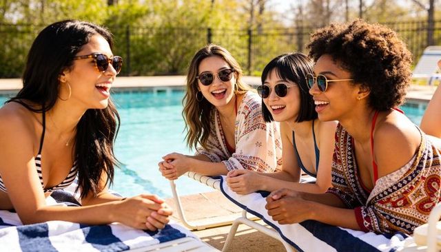 Laughing women lounging poolside in swimsuits and sunglasses enjoying sunny summer day