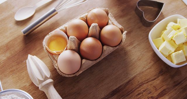 A carton of brown eggs, one cracked open, is placed on a wooden table alongside butter and baking utensils, with copy space. Fresh ingredients are prepared for a baking recipe, evoking a sense of home cooking and culinary preparation.