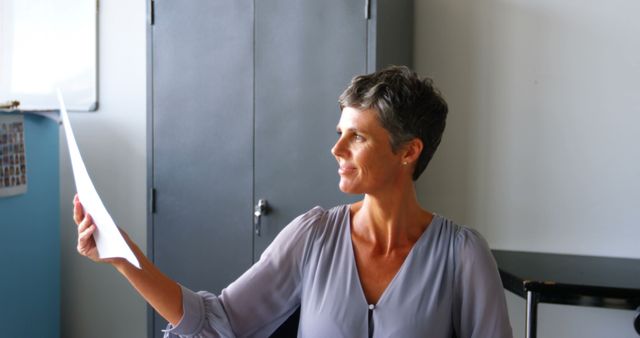 A middle-aged woman with short hair in a professional setting is reviewing a paper document in an office. She is wearing a blouse and seems to be engaged in her work. This image is suitable for business-related content, corporate websites, career guidance articles, and professional development resources.