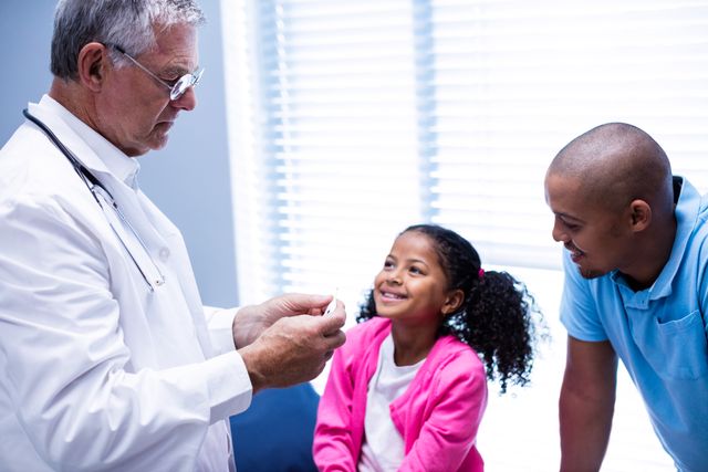 Doctor checking temperature of a smiling girl with her father present in a clinic. Ideal for use in healthcare, medical, pediatric, and family-related content. Can be used in articles, brochures, and websites promoting child health, medical services, and family care.
