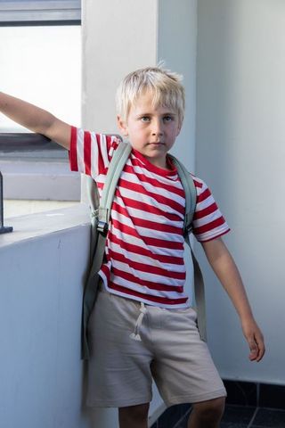 School-age boy leaning on window ledge in stairwell wearing striped t-shirt and backpack