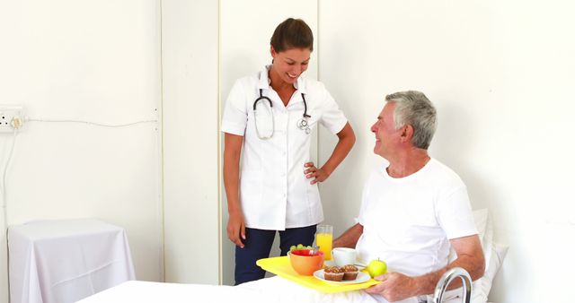 Nurse dressed in white uniform interacting with elderly male patient in hospital bed. Medical instruments hang from nurse's stethoscope. Patient looks pleased, holding food tray with healthy breakfast items including fruit, juice and bread. Could be used for healthcare, elderly care, medical services, hospital settings, or patient recovery themes.