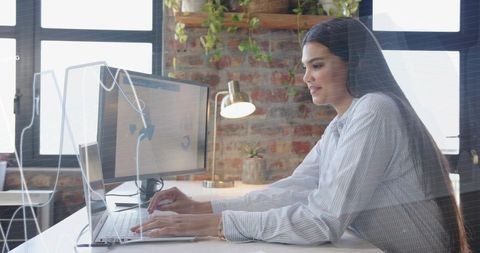 Mid-adult woman typing on laptop in modern loft workspace with exposed brick and greenery