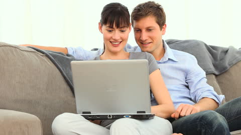Young Couple Smiling While Using Laptop on Sofa at Home