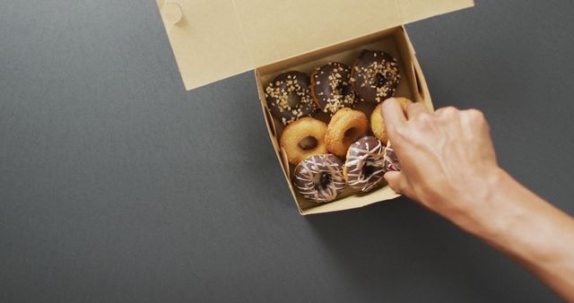 Hand reaching for a donut from a box containing an assortment of donuts with various toppings. Use this for themes around food, indulgence, dessert selections, or bakery aesthetics. Ideal for advertisements, food blogs, or marketing materials highlighting sweets and baked goods.