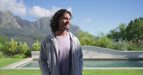 Smiling african american man standing by backyard pool with mountain view and casual hoodie