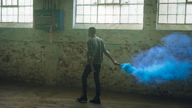 Rear view of a hip young biracial man in an empty warehouse, walking with a hand grenade with blue smoke.