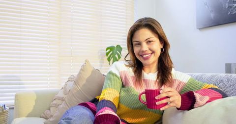 Cozy lifestyle portrait of woman smiling while holding magenta mug on sofa, colorful sweater