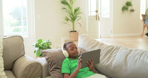 African American Boy Relaxing on Sofa Using Tablet with Headphones in Bright Living Room