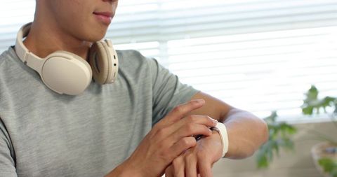 Asian man tapping smartwatch while wearing beige headphones and gray shirt, wearable tech