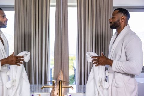 Mature african man in white bathrobe grooming at marble vanity with gold faucets
