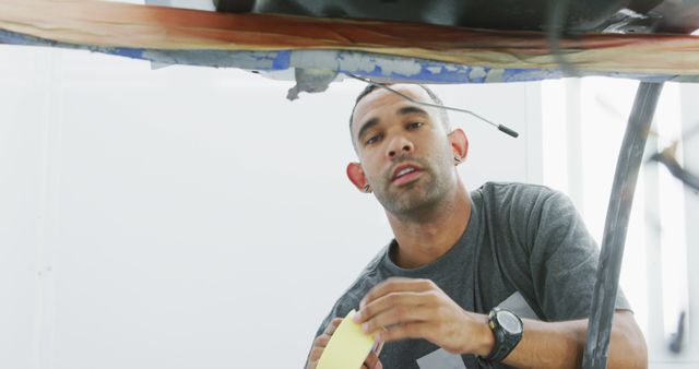 Male auto mechanic wearing grey t-shirt working meticulously under a vehicle. Image ideal for illustrating auto repair services, mechanical processes, maintenance tips, and professional auto garages. Useful for websites, brochures, training materials, and advertisements related to automotive repair and maintenance services.