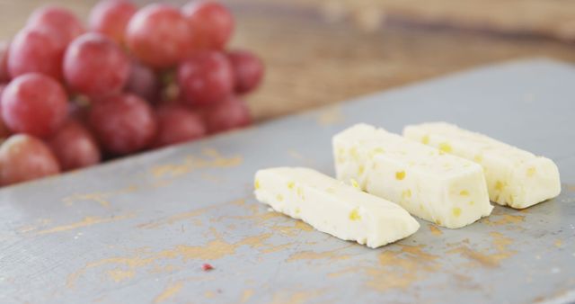 Slices of cheese with herbs are laid out on a cutting board, with a bunch of red grapes in the background, with copy space. A scene capturing a simple yet elegant snack setup, perfect for a wine pairing or a gourmet appetizer.