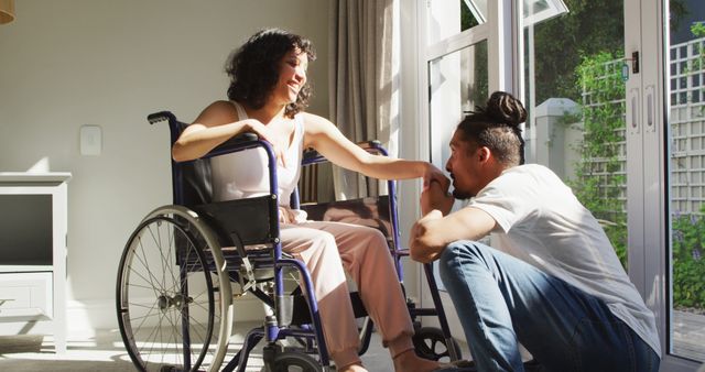 Happy biracial woman in wheelchair with smiling male partner kissing hand in sunny living room. wellbeing and domestic lifestyle with physical disability.