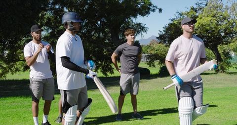 Preparing diverse cricket team adjusting pads and helmets holding bats on sunlit field