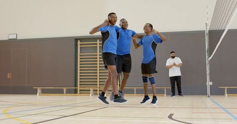 Diverse male volleyball teammates jumping and celebrating on indoor court with coach