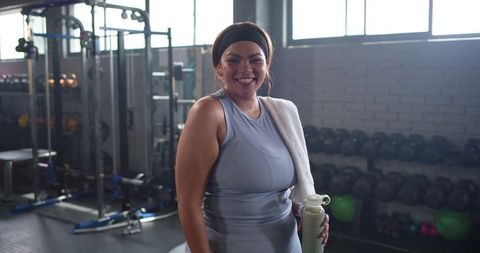 Confident woman finishing gym workout smiling with towel and bottle by dumbbells