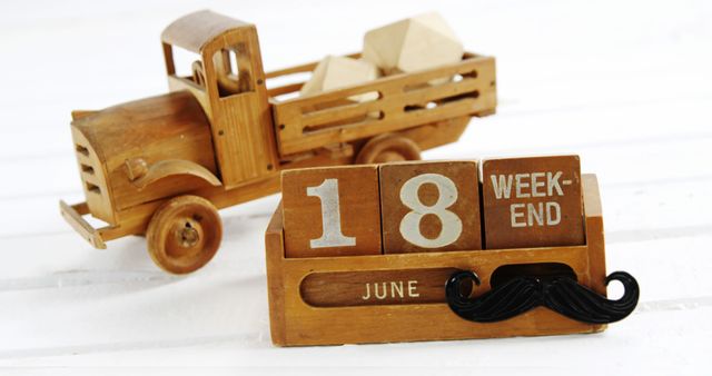A wooden calendar displaying the date June 18 with the word week-end sits next to a toy wooden truck, both on a white surface. The playful mustache on the calendar adds a whimsical touch to the scene, indicating a lighthearted approach to marking time.