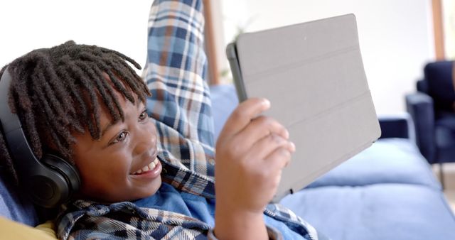 Young boy wearing headphones, using tablet while smiling on blue couch. Perfect for illustrating joy of technology, relaxed home environment, and modern childhood. Useful for education, tech product promotions, family blogs, or lifestyle articles.
