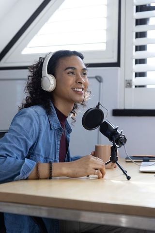 Non-binary podcaster speaking into desktop microphone in sunlit minimalist home studio