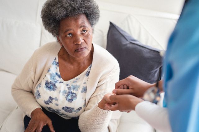 This image depicts a female doctor or nurse checking the fingers of an African American senior woman at home. It is ideal for use in articles or advertisements related to home healthcare, senior care services, medical checkups, and elderly support. It can also be used in brochures or websites promoting healthcare services, home visits by medical professionals, and patient care.