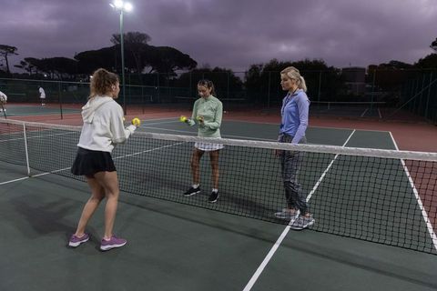 Women practicing tennis drills under floodlights on outdoor court at dusk