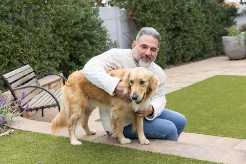 Mature man crouching in backyard holding golden retriever with tennis ball, pet moment