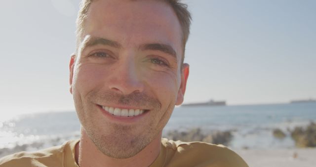 Young man smiling during a sunny day at the ocean with boats visible in the background. Ideal for travel brochures, vacation promotions, summer ads, and content emphasizing joy or relaxation by the seaside.