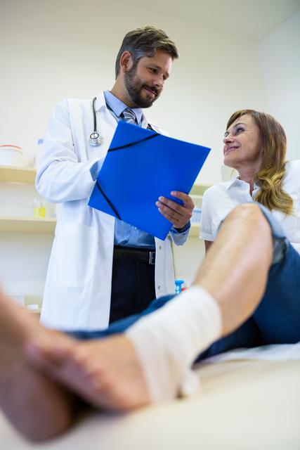 Doctor consulting a patient with a leg injury in a hospital setting. The patient has a bandaged leg and is smiling while the doctor reviews medical documents. This image can be used for healthcare websites, medical blogs, hospital brochures, and patient care articles.