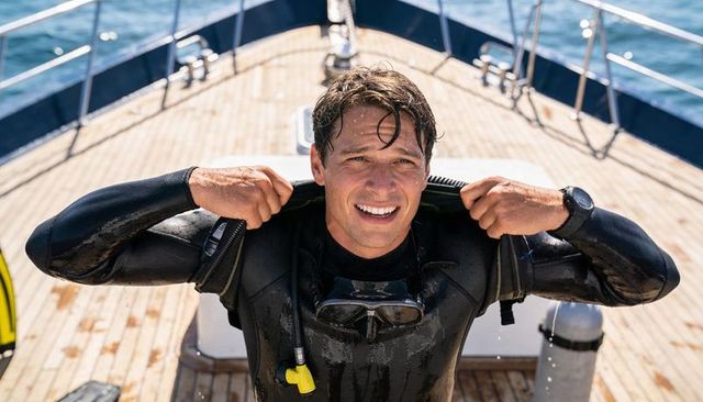 Smiling scuba diver adjusting wetsuit on sunlit boat deck with yellow regulator