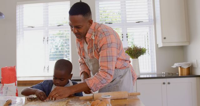 A father and his young son creating cookies in a modern, well-lit kitchen. The scene shows a warm bonding moment, with the father's guidance and patience evident as the son works carefully on the dough. This image is ideal for websites, blogs, and advertisements focused on family activities, parenting tips, and baking recipes. It captures the essence of family togetherness and the joys of teaching and learning new skills.