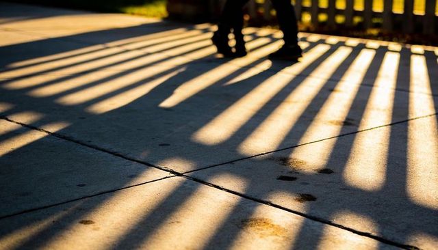 Golden-hour slatted fence casting striped shadows across sidewalk with walking boots