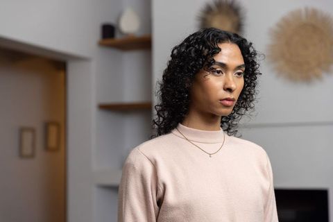 Non-binary model standing in minimalist living room wearing pale pink sweater and gold pendant