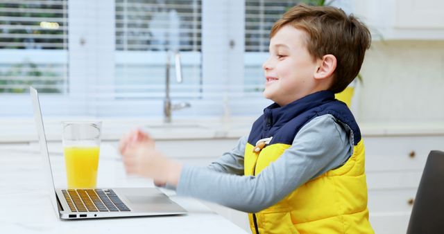 Young boy sitting in kitchen, using laptop for online education with glass of orange juice nearby, smiles while focusing on screen. Great for themes around home learning, children's education, modern technology, or healthy lifestyle for kids.