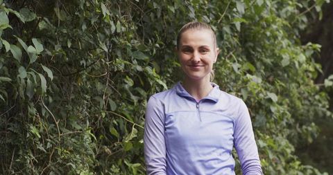 Smiling woman standing in park wearing light purple athletic top amid lush green foliage